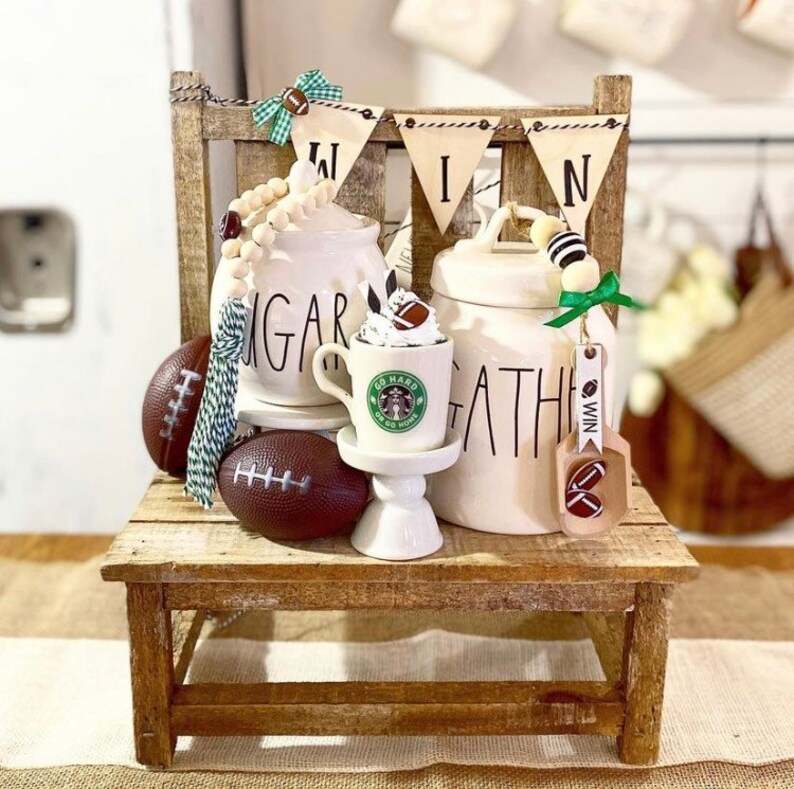 Op de afbeelding: Een houten bankje met een witte keramische suikerpot, een witte keramische koekpot en een klein wit keramisch standaard met een koffiekopje erop. De suikerpot zegt "Sugar" en de koekpot zegt "Gather". Er zijn voetballen, een groen-witte kwast en een houten bord met de tekst "Win" dat aan de koekpot hangt.