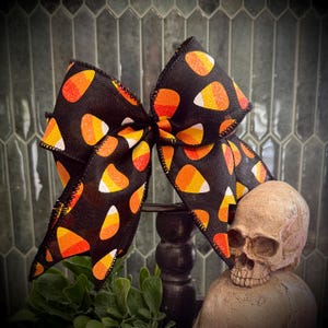 Puede incluir: Un gran lazo negro con un patrón de caramelos de maíz en naranja, amarillo y blanco. El lazo se coloca sobre una calavera decorativa y vegetación, creando un tema de Halloween.