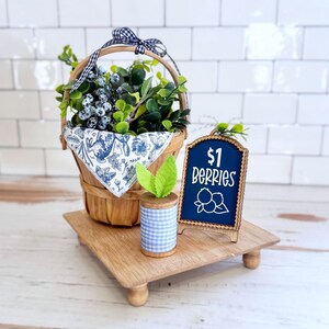 May include: A wooden basket with a blue and white patterned cloth, filled with green leaves and blue berries, tied with a gingham ribbon. Next to it is a wooden spool with light blue gingham ribbon and a small sign reading $1 BERRIES.