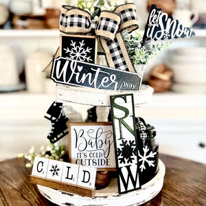 May include: A tiered tray decorated for winter with black and white buffalo check ribbon, a sign that says "Winter Way", a sign that says "Let It Snow", a sign that says "Baby It's Cold Outside", and a sign that says "Cold" with letter tiles.