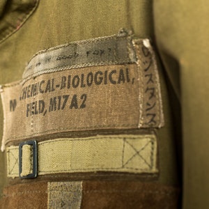 May include: A close-up of a green military jacket with a brown patch that reads "CHEMICAL-BIOLOGICAL, FIELD, MI7A2".
