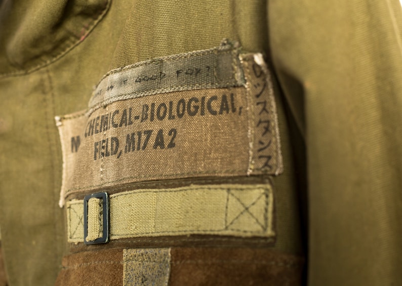 May include: A close-up of a green military jacket with a brown patch that reads "CHEMICAL-BIOLOGICAL, FIELD, MI7A2".