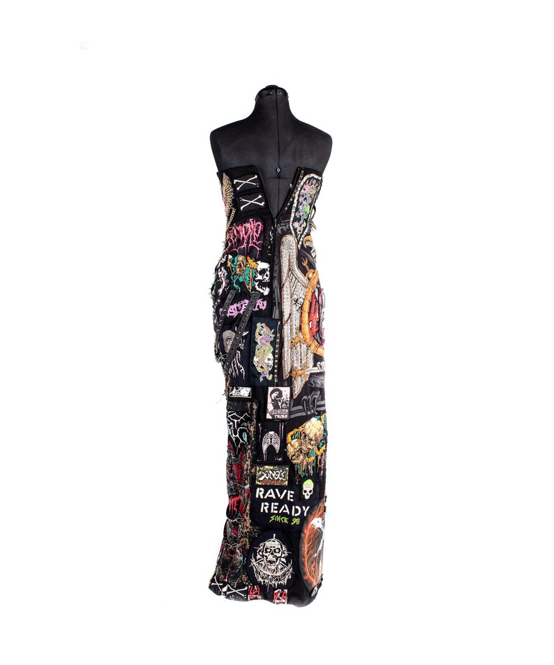 May include: A black strapless dress covered in colorful patches and embellishments. The patches feature various designs, including skulls, wings, and text. The dress is on a mannequin.