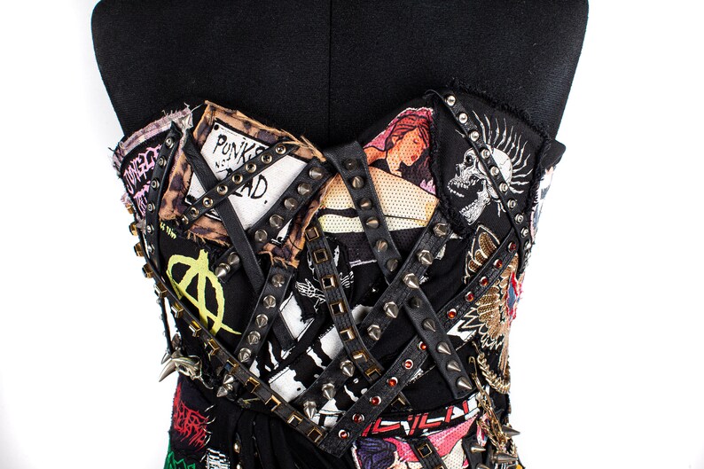 May include: A black strapless top with a punk rock aesthetic. The top is covered in patches, studs, and leather straps. Some of the patches include images of a skull, a woman, and the words "Punk's Not Dead."