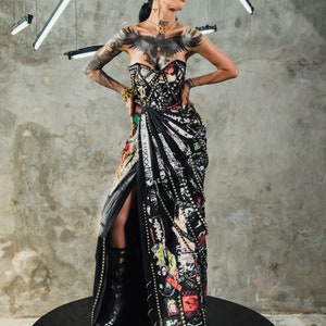 May include: A woman wearing a black, studded, and patched dress with a high slit. She has a black eagle tattoo on her chest and is wearing gold jewelry. The dress is adorned with safety pins, chains, and other embellishments. She is standing on a black platform.