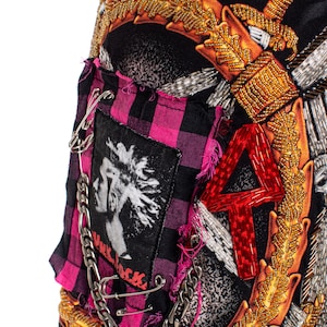 May include: A black fabric skirt with a pink and black plaid patch, a silver chain, safety pins, and a red beaded number "4". The skirt is decorated with gold and silver beaded embellishments, including a sword and a floral design.