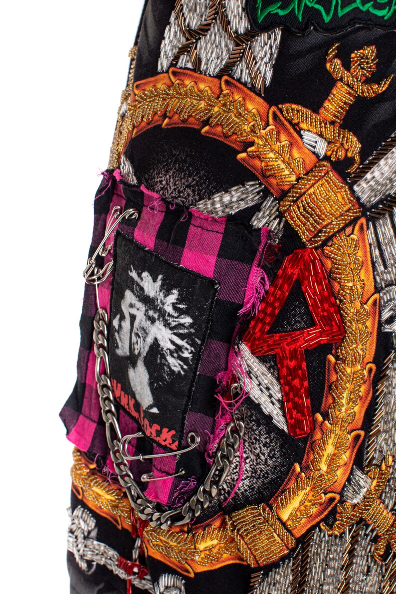 May include: A black fabric skirt with a pink and black plaid patch, a silver chain, safety pins, and a red beaded number "4". The skirt is decorated with gold and silver beaded embellishments, including a sword and a floral design.