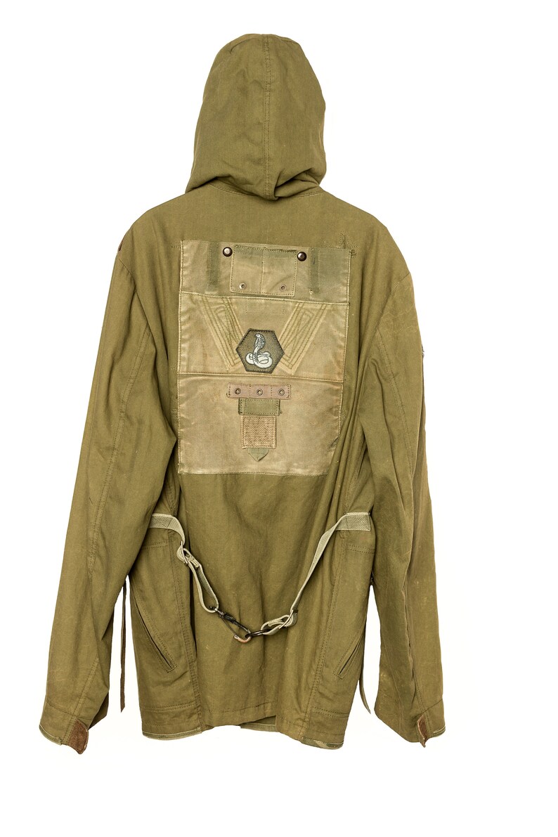 May include: A green hooded jacket with a large patch on the back. The patch features a snake design and a geometric pattern. The jacket has a belt and two pockets on the sides.
