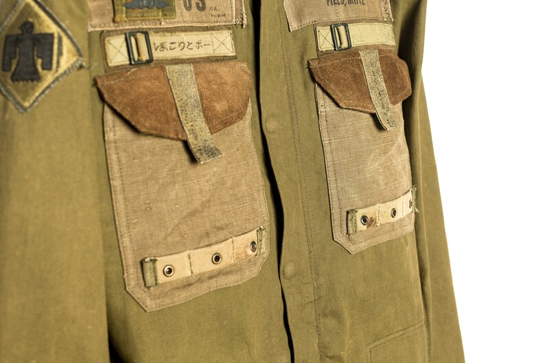 May include: A green utility shirt with multiple pockets and patches. The shirt has a large patch with a Native American design on the left side of the chest. The right side of the chest has a patch with the text "FIELD, WHITE" and a smaller patch with the text "US" and a graphic of a plane. The shirt also has a patch with Japanese text that reads "いんでこのコーナー".