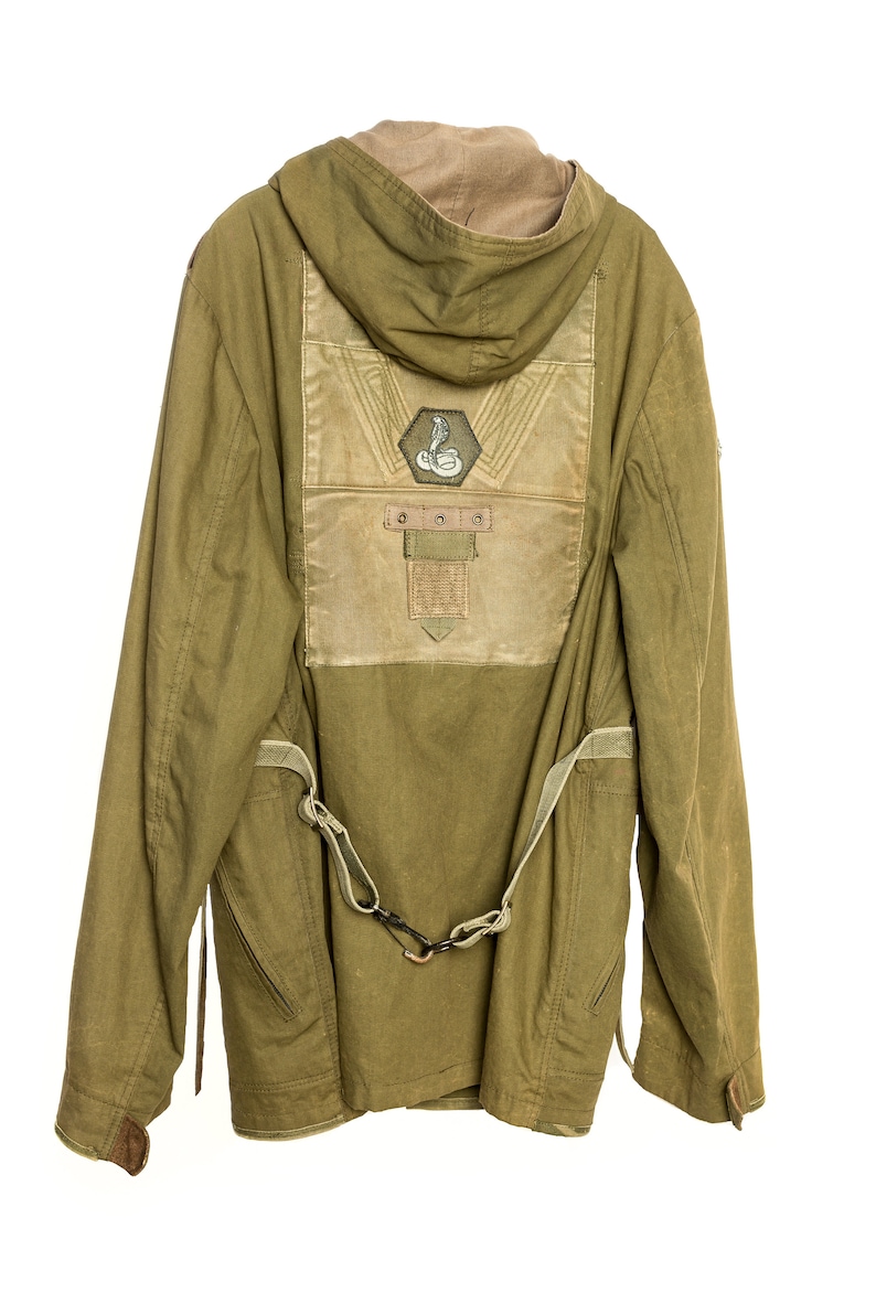 May include: A green hooded jacket with a brown patch on the back featuring a snake design and the text "DOOO". The jacket has a strap and buckle detail on the front.
