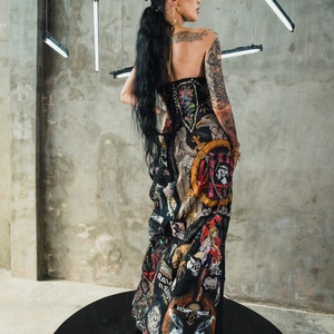 May include: A woman wearing a black dress with a variety of colorful patches and designs. The dress has a corset-style bodice and a flowing skirt. The woman has long black hair and is wearing a gold earring. The dress has a variety of patches with text, including "Rage Against The Machine", "Rave", and "Revolt".