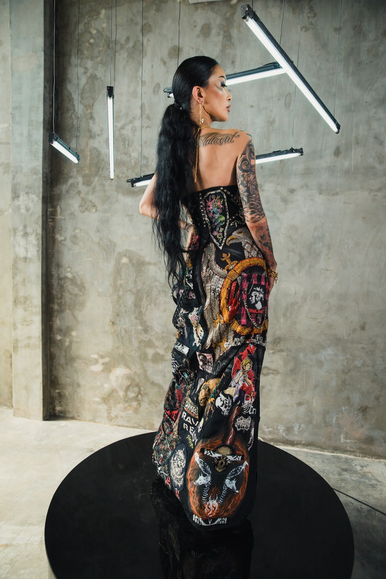 May include: A woman wearing a black dress with a variety of colorful patches and designs. The dress has a corset-style bodice and a flowing skirt. The woman has long black hair and is wearing a gold earring. The dress has a variety of patches with text, including "Rage Against The Machine", "Rave", and "Revolt".