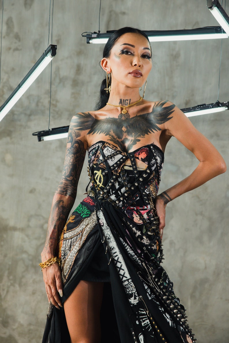 May include: A woman wearing a black corset with a punk rock aesthetic. The corset is covered in studs, safety pins, and patches. She has a large black bird tattoo on her chest and the word "AIGI" tattooed on her neck.