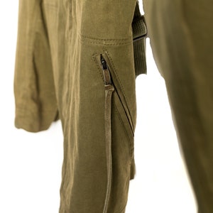 May include: A close-up of a green jacket with a zipped pocket and a brown leather strap. The jacket is made of a durable, canvas-like material.