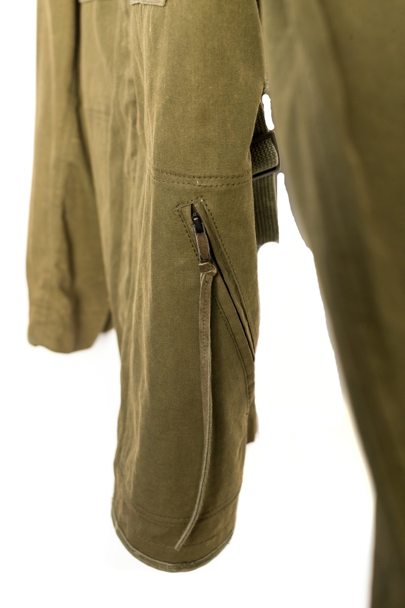 May include: A close-up of a green jacket with a zipped pocket and a brown leather strap. The jacket is made of a durable, canvas-like material.