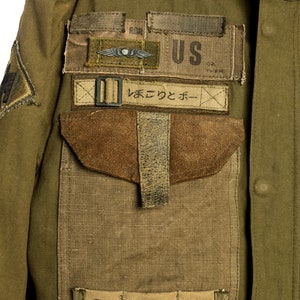 May include: A green military jacket with a large patch pocket on the left side. The pocket has a brown suede flap and a strap with a buckle. There are several patches sewn onto the jacket, including a US flag patch, a patch with a winged insignia, and a patch with Japanese text that reads "まえて つと ボー".