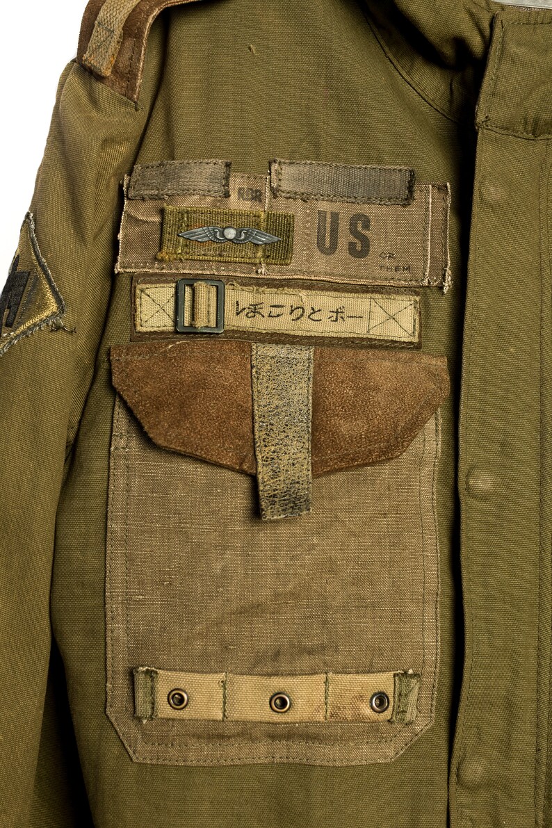 May include: A green military jacket with a large patch pocket on the left side. The pocket has a brown suede flap and a strap with a buckle. There are several patches sewn onto the jacket, including a US flag patch, a patch with a winged insignia, and a patch with Japanese text that reads "まえて つと ボー".