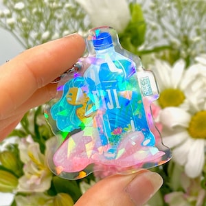 May include: A holographic charm featuring a mermaid holding a blue Fiji water bottle. The charm is pink and iridescent with glitter accents.