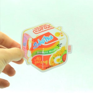 May include: A sticker of a cartoon illustration of a box of Indomie Mi Goreng instant noodles. The box is yellow, orange, and green with the text "Indomie" and "Mi Goreng" in a stylized font. The sticker is white with a light blue border.