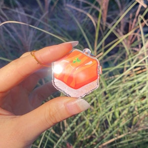 Pu&ograve; includere: Un ciondolo in acrilico trasparente con un'illustrazione colorata di un pezzo di sushi. Il sushi ha strati arancioni e bianchi, con dettagli verdi. Il ciondolo &egrave; appeso a una catena color argento.