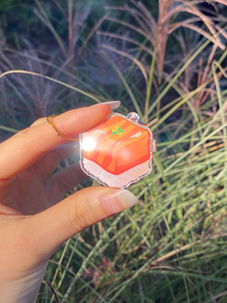 Pu&ograve; includere: Un ciondolo in acrilico trasparente con un'illustrazione colorata di un pezzo di sushi. Il sushi ha strati arancioni e bianchi, con dettagli verdi. Il ciondolo &egrave; appeso a una catena color argento.