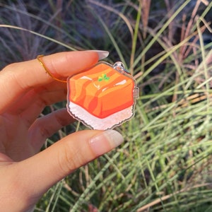Pu&ograve; includere: Un ciondolo in acrilico trasparente con un'illustrazione colorata di un dolce, forse baklava. Il ciondolo ha una catena d'oro e una piccola chiusura. Il dolce &egrave; arancione e bianco con una guarnizione verde.