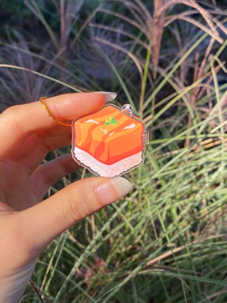 Pu&ograve; includere: Un ciondolo in acrilico trasparente con un'illustrazione colorata di un dolce, forse baklava. Il ciondolo ha una catena d'oro e una piccola chiusura. Il dolce &egrave; arancione e bianco con una guarnizione verde.