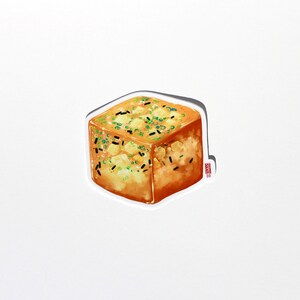 May include: A sticker featuring a square of golden-brown food, possibly tofu or a similar dish. It is garnished with green onions and black sesame seeds. The sticker has a white border and a small logo in the bottom right corner.