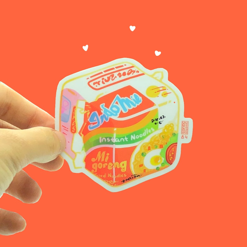 May include: A sticker of a red, yellow, and green Indomie Mi Goreng instant noodle package. The package has the text "Indomie" and "Mi goreng" on it. The sticker is on a bright orange background.
