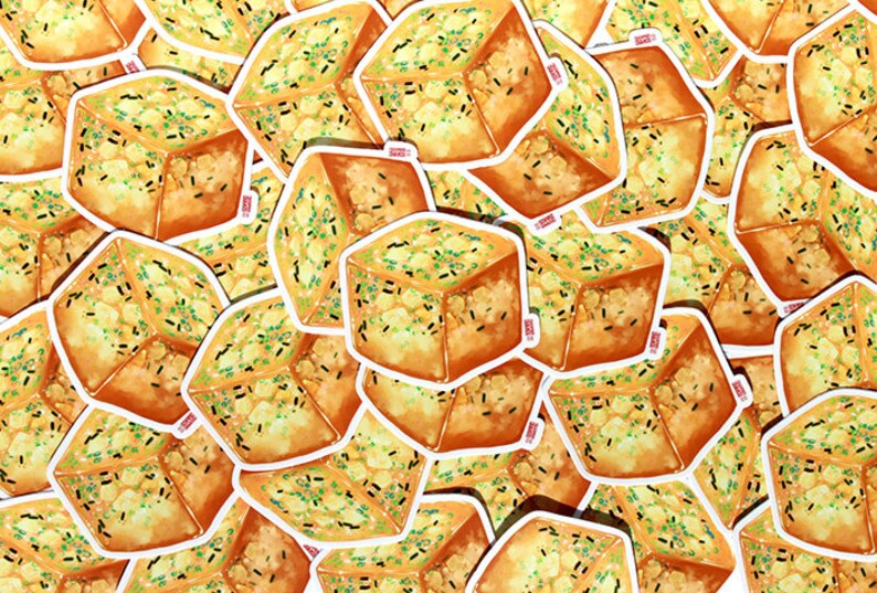 May include: A collection of stickers featuring a watercolor illustration of a cube-shaped food item. The food has a golden-brown exterior, a light yellow interior, and is sprinkled with green and black specks. The stickers have a white border.