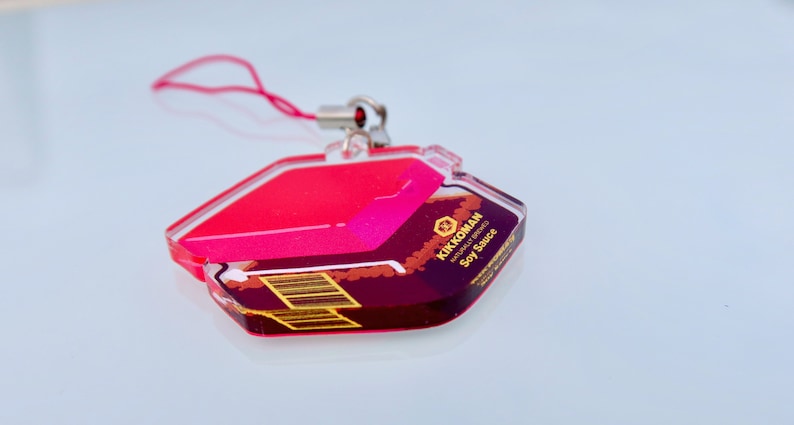 May include: A red and gold acrylic keychain shaped like a bottle of Kikkoman Naturally Brewed Soy Sauce. The keychain has a red string attached to it.