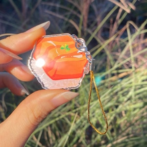 Pu&ograve; includere: Un ciondolo trasparente in acrilico con un design colorato. Il ciondolo presenta uno sfondo arancione brillante con un quadrifoglio verde. Ha un fermaglio in metallo color argento e una catena color oro. Il ciondolo &egrave; tenuto davanti a uno sfondo verde sfocato.