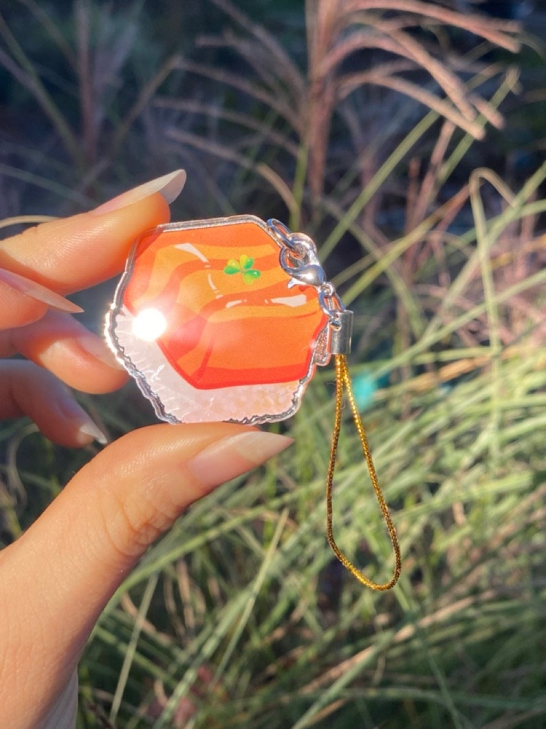 Pu&ograve; includere: Un ciondolo trasparente in acrilico con un design colorato. Il ciondolo presenta uno sfondo arancione brillante con un quadrifoglio verde. Ha un fermaglio in metallo color argento e una catena color oro. Il ciondolo &egrave; tenuto davanti a uno sfondo verde sfocato.