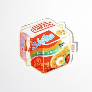 May include: A sticker of a red and yellow Indomie Mi Goreng instant noodle package with the text "Indomie" and "Mi goreng" in white. The package has a picture of noodles with an egg, peas, and a tomato.
