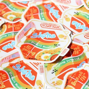 May include: A pile of colorful stickers featuring the Indomie brand of instant noodles. The stickers are designed to look like the Indomie packaging, with the brand name and product name in a stylized font. The stickers are a fun and quirky way to show your love for Indomie.