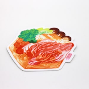 May include: A sticker featuring a colorful illustration of a Japanese hot pot with meat, vegetables, and noodles. The sticker has a white background and a pink stamp with Japanese text.