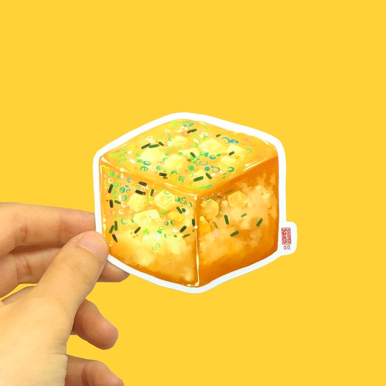 May include: A sticker featuring a square-shaped illustration of a golden-brown, savory food item, possibly a type of bread or pastry, topped with green and yellow garnishes. The sticker is held against a yellow background.