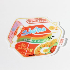 May include: A sticker of a red and yellow Indomie Mi Goreng instant noodle package with a bowl of noodles, an egg, and a tomato. The text on the package reads "Indomie Instant Noodles Mi Goreng Fried Noodles".