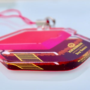 May include: A pink and black acrylic keychain with the text "KIKUCHI KAN" and "Soy Sauce" printed on it. The keychain is shaped like a hexagon and has a silver ring.