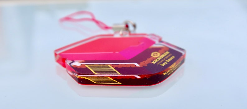 May include: A pink and black acrylic keychain with the text "KIKUCHI KAN" and "Soy Sauce" printed on it. The keychain is shaped like a hexagon and has a silver ring.