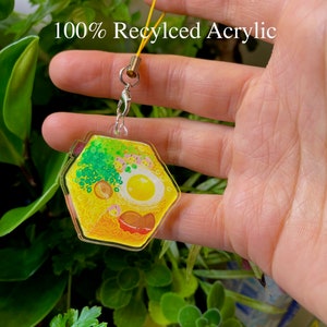 May include: A yellow and green hexagonal keychain with a cartoon illustration of a bowl of ramen noodles. The keychain is made of 100% recycled acrylic.