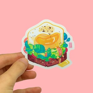 Könnte beinhalten: Ein Aufkleber mit einer Cartoon-Illustration eines Frühstücks-Sandwiches mit Spiegelei, Avocado und grünem Blattgemüse auf einem gerösteten Brötchen. Der Aufkleber hat einen weißen Hintergrund und einen rosa Rand.