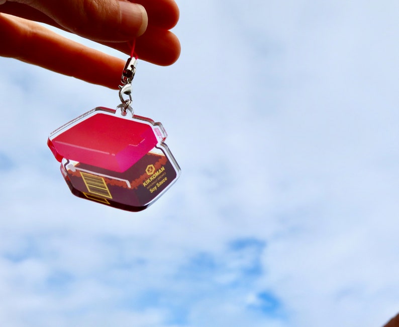 May include: A clear acrylic keychain shaped like a box of Kikkoman soy sauce. The box is open and the lid is red. The keychain has a silver metal ring.