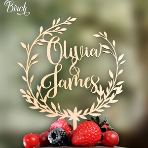May include: A wooden cake topper with a leafy wreath design and the names "Olivia & James" written in cursive. The topper is placed on a chocolate and white cake with red and black berries.