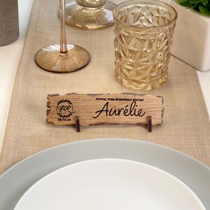 May include: A rustic wooden place card with the name "Aur&eacute;lie" engraved on it. The card also features a monogram and the date "08.10.24". The card is sitting on a table with a glass of water and a plate.