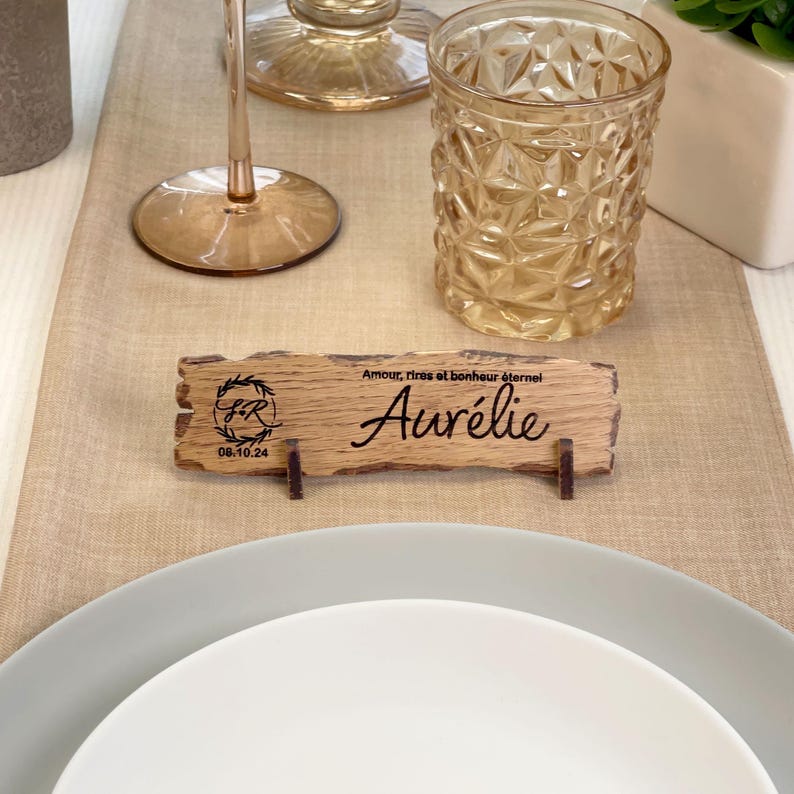 May include: A rustic wooden place card with the name "Aur&eacute;lie" engraved on it. The card also features a monogram and the date "08.10.24". The card is sitting on a table with a glass of water and a plate.