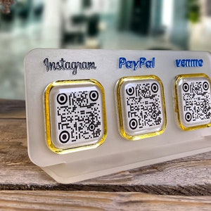 May include: A white acrylic stand with three gold-rimmed QR code stickers. The stickers are labeled "Instagram", "PayPal", and "Venmo".