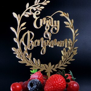 May include: Gold cake topper with a leafy wreath design and the names "Emily & Benjamin" in cursive script. The topper is placed on a chocolate cake decorated with fresh berries.