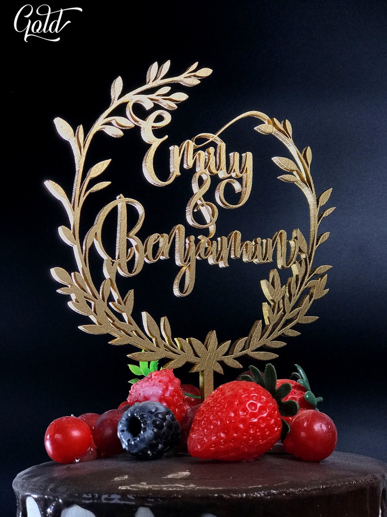May include: Gold cake topper with a leafy wreath design and the names "Emily & Benjamin" in cursive script. The topper is placed on a chocolate cake decorated with fresh berries.
