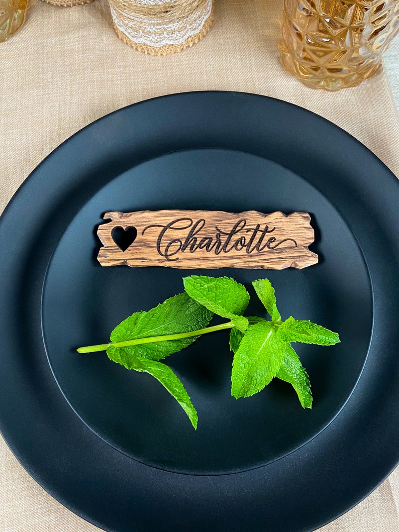 May include: A wooden place card with a heart cutout and the name "Charlotte" engraved on it. The place card is on a black plate with a sprig of green mint.